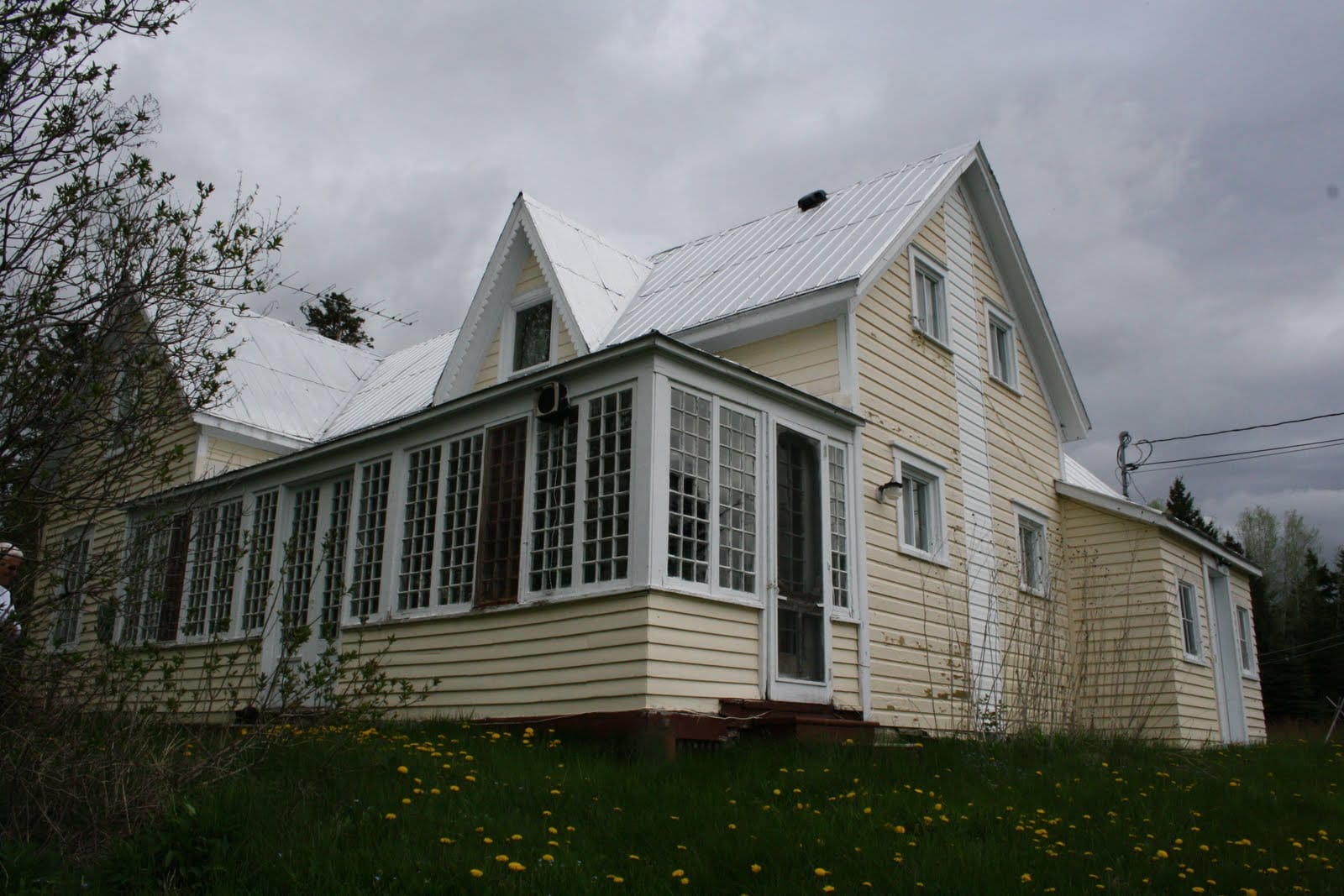 A large, old coastal farmhouse, covered in pale yellow clapboard, with a sharply peaked metal roof and a large porch covered in windows.